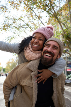 Vertical portrait of young cheerful multiracial happy couple in love hugging and smiling in piggyback pointing somewhere outdoors street. Smiling people dressed in winter clothes having fun together