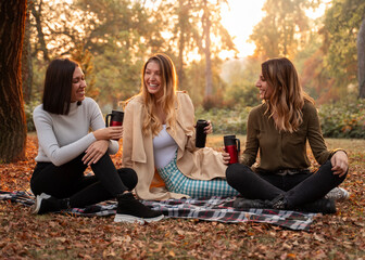 Three young adult beautiful charismatic woman sitting on blanket in park, drinking coffee and...