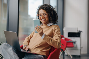 Stylish business woman is use phone while working on laptop in modern office near window