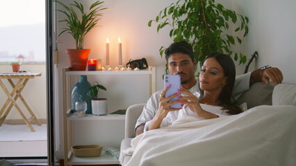 Married couple watching smartphone at living room closeup. Husband hugging wife