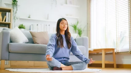  Happy young Asian woman practicing yoga and meditation at home sitting on floor in living room in lotus position and relaxing with closed eyes. Mindful meditation and wellbeing concept © Monster Ztudio