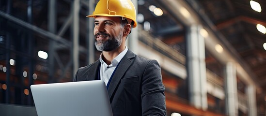 Handsome man in modern industrial environment, holding laptop, walks in factory with a hard hat.