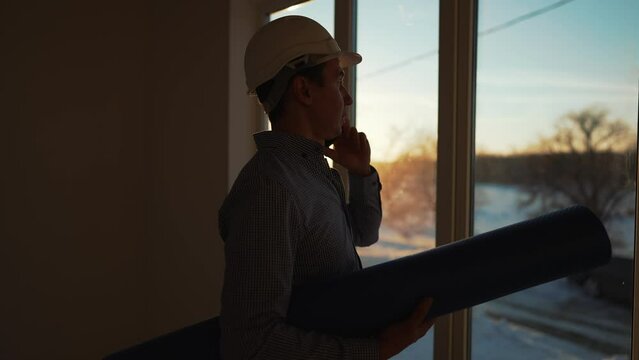 Male builder in hard hat stands at window, silhouette. Business concept of an engineer at construction site, business plan. Construction engineer on phone speaks about concept of business idea.