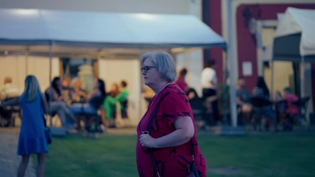 Panning shot of a white Woman with Glasses wearing red T-shirt walking around historical City Bardejov during a blue hour at night in slow motion, surrounded by people and lights.