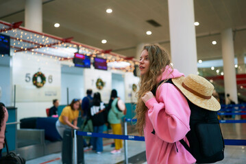 Holiday traveler at airport terminal, backpack and hat in hand, Christmas decor adorns the space...