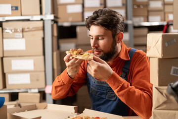 man in work clothes, a warehouse worker eats a meat pizza from fast food for lunch while sitting at...