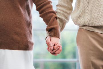 Close-up shot of a couple holding hands with interlocking fingers with blurred green windows in the...