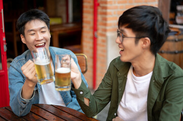 Two happy young Asian male friends are cheering and enjoying beers at a restaurant or bar together.