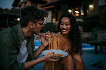 A multiethnic couple share pasta and laughter outdoors - obrazy, fototapety, plakaty