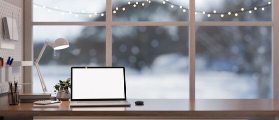 Home office workspace with a white-screen laptop on a wooden desk against the window on winter day.