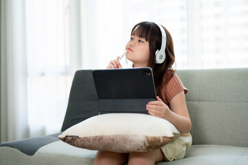 An adorable young Asian girl studying online at home on her digital tablet while sitting on a sofa.