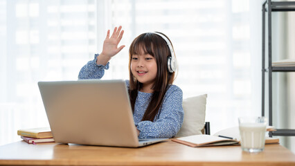 A cheerful and cute young Asian girl is raising her hand to answer a question while studying online.