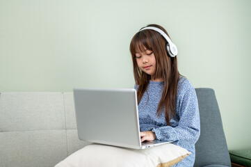 A cute young Asian girl wearing headphones is studying online on a laptop while sitting on a sofa.
