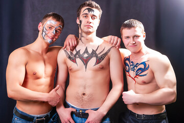 Fototapeta na wymiar Group of Three Masculine Caucasian Brunette Men Painted With Unique Face and Body Art Painting Posing Together Connected Against Black