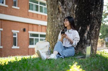 A positive Asian female college student is using her smartphone while relaxing under the tree.