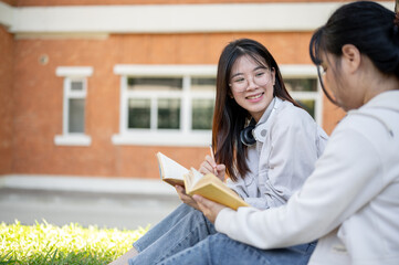 Obraz premium A female student is reading a book while relaxing under the tree in a campus park with her friend.