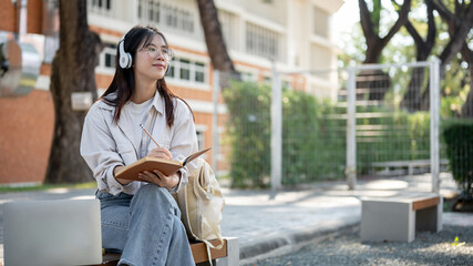 A positive female is listening to music and keeping her diary while resting on a bench in a park.