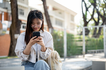Fototapeta premium A positive Asian woman is chatting with her friends on her phone while resting on a bench in a park.