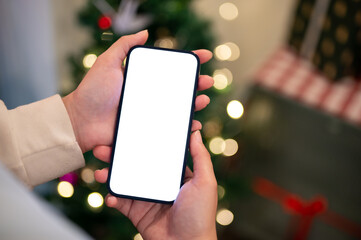 A woman holding a phone mockup over a blurred background of a Christmas tree and gifts in the room.