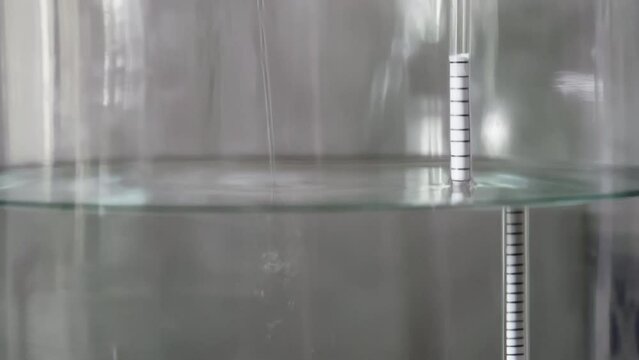 alcohol distilling process closeup - liquid distilled transparent liquid flowing in large glass jar with floating alcoholometer in it.