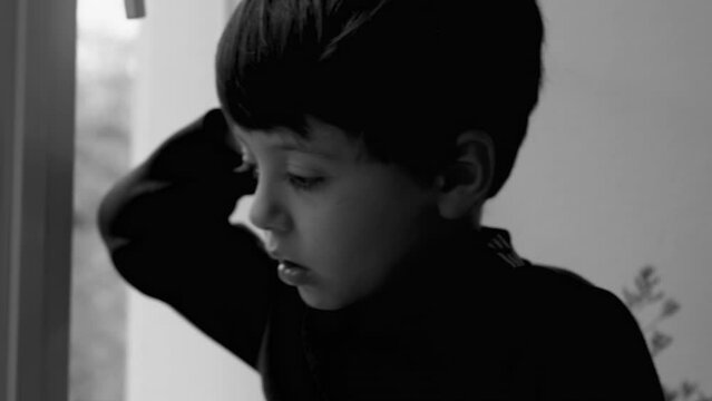 Child Seated by Window Feeling Depressed and Lonely in dramatic black and white. Little boy struggles with mental illness leaning on glass, moody gloomy feeling