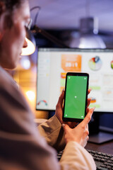 Entrepreneur looking at green screen mock up chroma key smartphone with isolated display, analyzing marketinf graphs report. Executive manager working late at night in startup office
