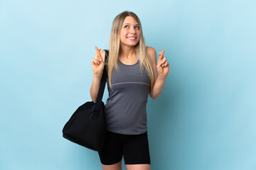 Young sport woman with sport bag isolated on blue background with fingers crossing and wishing the...