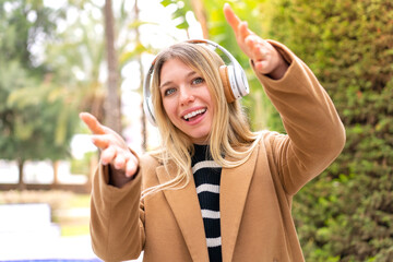 Young pretty blonde woman listening music with headphones at outdoors presenting and inviting to come with hand