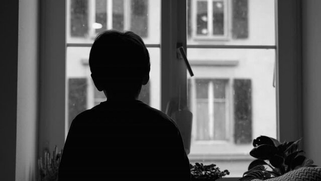Back of sad depressed child staring at snow fall from apartment's window. Pensive young boy stuck at home, struggling with depression in monochrome, black and white