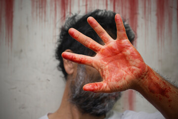 A middle aged man with a messy beard and blood-stained hands covering his face stood in front of a wall smeared with blood stains, concept of crime and murder