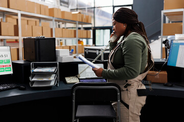 Stockroom african american worker holding clipboard, analyzing goods inventory report in storehouse. Storage room employee talking at landline phone with remote customer discussing delivery details