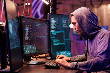 Asian criminal in hood hacking server and stealing data while committing online crime. Young hacker running illegal malicious software code on computer screen for cracking password - obrazy, fototapety, plakaty