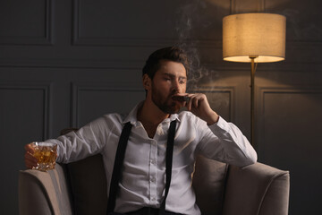 Handsome man with glass of whiskey smoking cigar in armchair at home