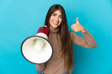 Young caucasian woman isolated on blue background holding a megaphone with thumb up - obrazy, fototapety, plakaty