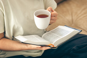 Woman holding a cup of tea and reading a book on the sofa at home, close up.