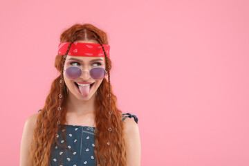 Stylish young hippie woman in sunglasses showing her tongue on pink background, space for text