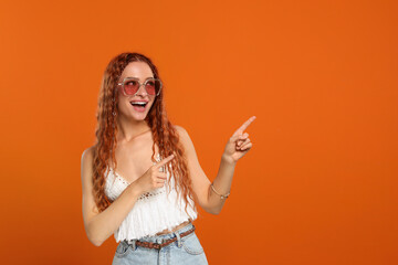 Stylish young hippie woman in sunglasses pointing at something on orange background, space for text