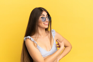 Young caucasian woman in swimsuit in summer holidays isolated on yellow background pointing back