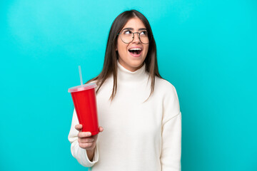 Young caucasian woman drinking soda isolated on blue background looking up and with surprised...