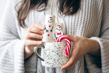 Cup of hot drink with marshmallow snowmen and candy in female hands.