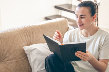 Young Caucasian woman reading a book, sitting on the sofa at home.