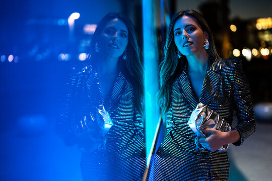 Woman holding clutch bag and standing near glass wall with neon lighting