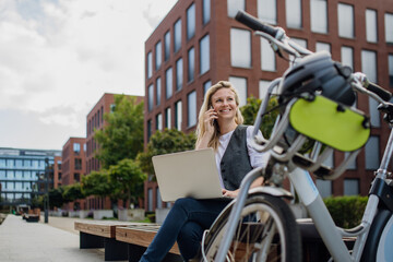 Businesswoman, freelancer or manager working outdoors in city park. Woman with laptop on knees making call on smartphone, sitting on park bench during sunny day. Concept of working remotely.