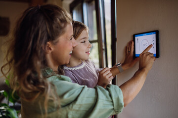 Girl helping mother to adjust, lower heating temperature on thermostat. Concept of sustainable, efficient, and smart technology in home heating and thermostats. - obrazy, fototapety, plakaty