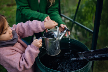 Girl and mother scooping rainwater from barell under downspout. Concept of water conservation in...