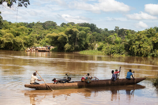 Transport of people across the Mbari river by locally made canoes and by ferry boat. Passengers colorfully dressed, transporting variety of goods purchased in local market on weekend day