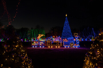 Landscape view of colorful holiday lights at night, featuring a festive toy train and cars design, with a blue Christmas tree in background
