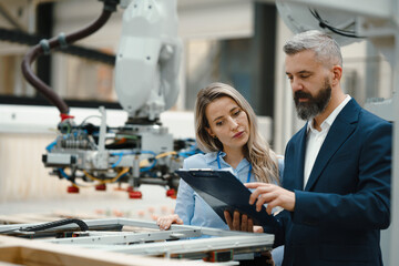 Female engineer and male production manager standing in modern industrial factory, talking about production. Manufacturing facility with robotics and automation. - obrazy, fototapety, plakaty