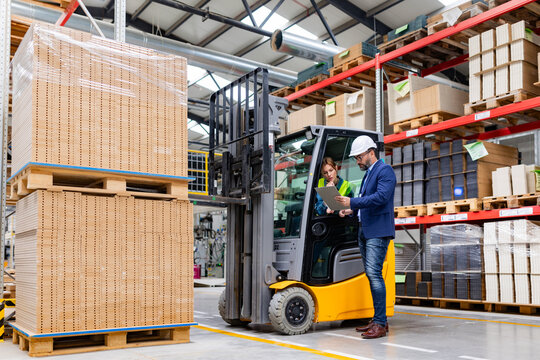 Forklift driver talking with manager in modern industrial factory. Woman warehouse worker discussing order, order picking. Warehouse worker preparing products for shipmennt, delivery, checking stock