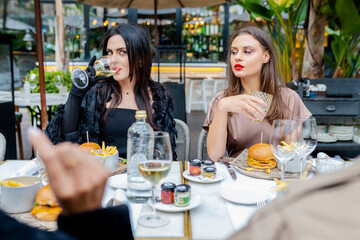 Group of multiracial stylish friends having lunch, drinking wine and eating burguers sitting on the terrace of a restaurant outdoors.